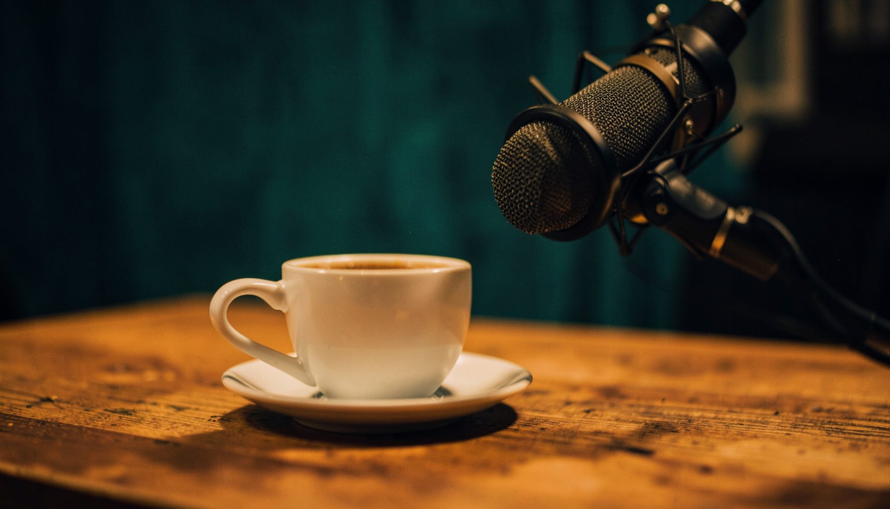 A warm, softly lit coffee cup on a wooden table beside a microphone, with deep teal and amber tones and a cozy, intimate podcast-studio mood.